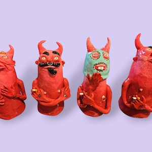 May include: Four red ceramic devil figurines with horns, each displaying distinct facial expressions and hand gestures. One figure wears a light blue mask, while others feature details like a nose ring, moustache, and small cigarettes held in their hands.