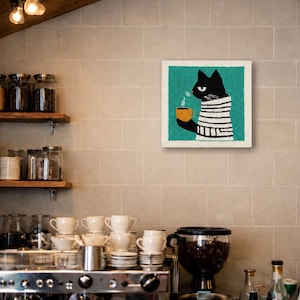 May include: A square punch needle wall art piece featuring a black cat wearing a white and black striped sweater, holding a steaming yellow mug against a teal background. The artwork is displayed on a tiled wall above a coffee station.