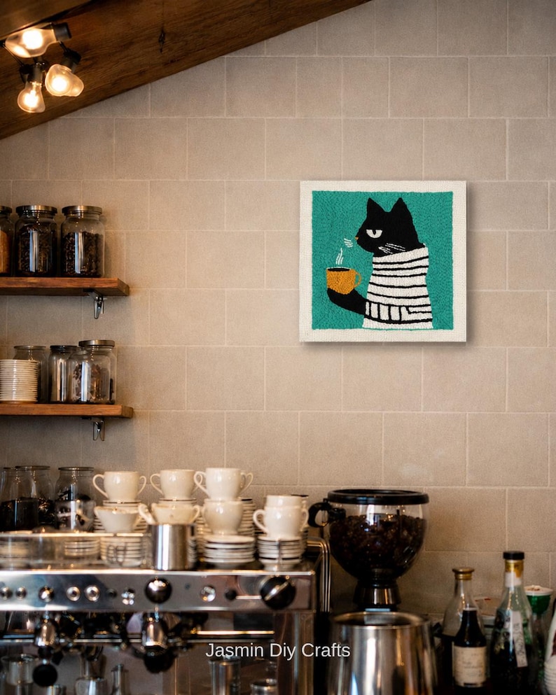 May include: A square punch needle wall art piece featuring a black cat wearing a white and black striped sweater, holding a steaming yellow mug against a teal background. The artwork is displayed on a tiled wall above a coffee station.