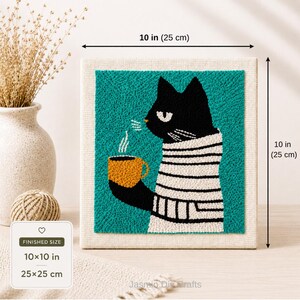 May include: A square punch needle embroidery art piece featuring a black cat wearing a white and black striped sweater, holding a yellow mug with steam rising. The artwork measures 10 inches by 10 inches and is set against a cream-colored fabric.