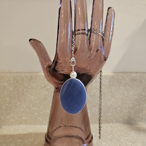 May include: A silver chain necklace with a blue oval pendant and a white pearl accent. The necklace is displayed on a purple glass hand-shaped jewelry holder. The background is a neutral speckled surface.