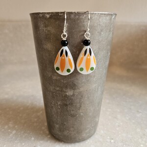 May include: A pair of teardrop-shaped earrings featuring a white base with orange petal-like patterns, black accents, and small green dots. Each earring is suspended from a silver-toned hook by a small black bead and a twisted silver wire link.