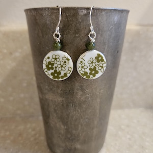 Puede incluir: Pendientes colgantes con colgantes circulares blancos con un diseño floral verde. Cada pendiente tiene una pequeña cuenta verde y un gancho plateado. Los pendientes se muestran sobre un fondo gris.