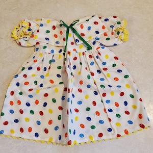 May include: White baby dress with a colorful polka dot pattern. The dress features a Peter Pan collar, short sleeves with yellow trim, and a green ribbon bow. The hemline is trimmed with yellow scalloped edging.
