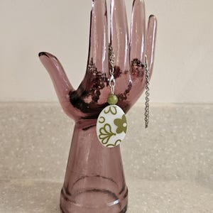 Könnte beinhalten: Ohrringe mit einem weißen, ovalen Anhänger mit einem grünen Blumenmuster. Die Ohrringe hängen an einer silbernen Kette und werden auf einem lila gläsernen, handförmigen Ständer präsentiert.