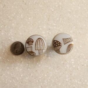 May include: Two white and gold mushroom-themed cabinet knobs. The knobs feature a circular design with various mushroom illustrations in gold. A U.S. quarter is next to the knobs for size comparison. The knobs are likely for furniture or decorative use.