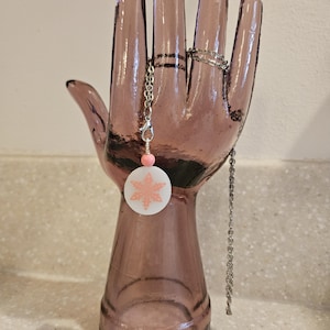 May include: A clear, mauve-colored glass hand sculpture holds a silver chain necklace with a white pendant featuring a pink snowflake design. A silver ring is on the middle finger, and a pink bead accents the necklace.