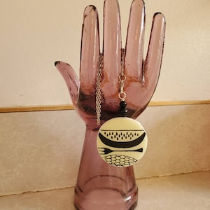 May include: A white ceramic pendant necklace with a black and white design of a watermelon, corn, and a fork. The necklace is hanging on a glass hand-shaped display stand.