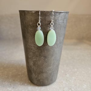 May include: A pair of dangle earrings featuring smooth, oval-shaped light green beads suspended from silver-toned metal hooks. Each earring includes a small, clear, faceted accent bead positioned directly above the larger green oval component.