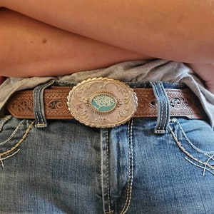Könnte beinhalten: Ein brauner Ledergürtel mit einer silbernen Schnalle mit einem türkisfarbenen Design. Der Gürtel wird um die Taille einer Person getragen, die eine blaue Jeans trägt.