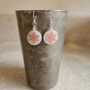 May include: A pair of dangle earrings featuring circular white discs with a coral-colored snowflake pattern. The discs are suspended from silver-toned metal hooks, each accented with a small, clear, faceted bead above the disc attachment point.