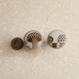 May include: Two round cabinet knobs with a mushroom design. The knobs have a white background with a brown mushroom illustration. The knobs are next to a quarter for size comparison. The knobs have a silver metal rim.