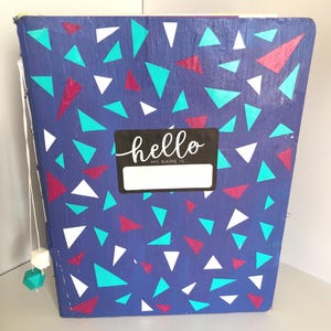Può includere: Un quaderno blu con un motivo geometrico di triangoli bianchi, turchese e bordeaux. Un'etichetta nera con la parola "hello" e uno spazio per un nome è sul davanti. Un filo con perline decorative pende dal lato.