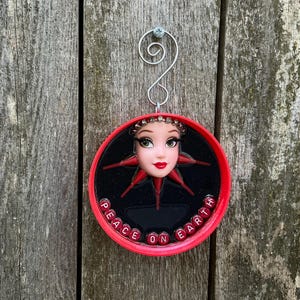 May include: A round, red Christmas ornament with a doll face at the center, surrounded by a red starburst design on a black background. The words "PEACE ON EARTH" are spelled out in red beads around the edge. A silver wire loop is attached for hanging.
