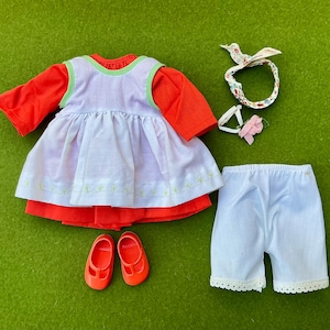 May include: A doll's outfit set on a green surface. The set includes a red long-sleeved shirt, a white apron with green trim, white shorts with lace trim, red shoes, and a floral headband with a pink flower.