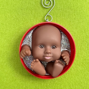 May include: A baby doll ornament with a red circular frame. The baby has brown skin, brown eyes, and a smiling expression. The background is silver glitter. A silver wire is attached for hanging.