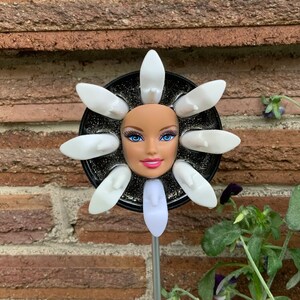 May include: A whimsical flower sculpture featuring a doll's head with blue eyes and pink lips at the center. White shoe-shaped petals surround the face, set against a black, glittery background. The sculpture is mounted on a silver metal stake.