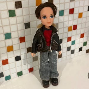 May include: A Bratz doll of a young man with brown hair and blue eyes. He wears a black leather jacket with a faux fur collar over a red shirt, distressed denim jeans, and black boots. The doll is standing in front of a tiled wall.