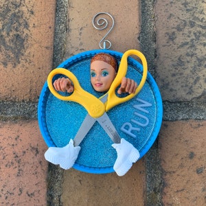 May include: A whimsical ornament featuring a doll head, yellow scissors, and white shoes, set against a blue glitter background. The word "RUN" is displayed. The ornament has a silver wire for hanging.