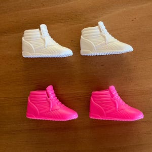 May include: Four miniature high-top sneakers. Two are white, and two are bright pink. The shoes have a ribbed design on the sides and a textured sole. The sneakers are arranged in a square on a wooden surface.