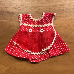 May include: A red and white polka dot doll dress with white trim and two white buttons. The dress has a ruffled skirt and a scalloped trim around the neckline and sleeves. The dress is made of a lightweight fabric.