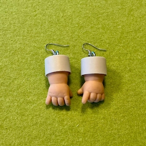 May include: A pair of novelty earrings featuring miniature baby doll hands. The hands are flesh-toned with a white band at the wrist, attached to silver-colored ear wires. The earrings are set against a green background.