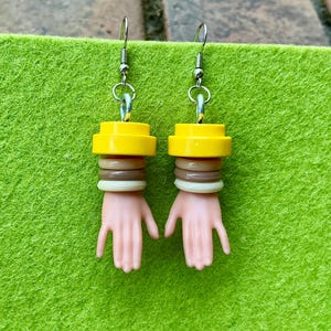 May include: A pair of dangle earrings featuring miniature plastic hands in a light skin tone. Each earring has a yellow cylindrical top piece, followed by a stack of three small rings in white, brown, and cream colors, connected to a silver hook.