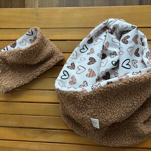 Könnte beinhalten: Zwei braune Fleece-Halswärmer mit einem weißen Stofffutter. Das Futter hat ein Muster aus rosa, braunen und schwarzen Herzen.