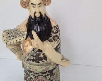 Op de afbeelding: Een wit, crème en gouden beeldje van een Chinese godheid met een zwarte baard en een rode, groene en gouden gepatroneerde robe. Het beeldje staat op een wit oppervlak.