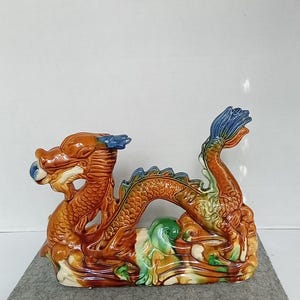 Op de afbeelding: Een keramiek drakenbeeldje met een bruine, groene en blauwe glazuur. De draak is opgerold met zijn staart in zijn bek.