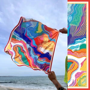 Op de afbeelding: Een kleurrijke zijden sjaal met een levendig abstract ontwerp, met tinten blauw, rood, oranje en geel. De sjaal heeft een rode rand en wordt omhoog gehouden tegen een blauwe lucht en oceaan achtergrond. Het ontwerp bevat golvende lijnen en organische vormen.