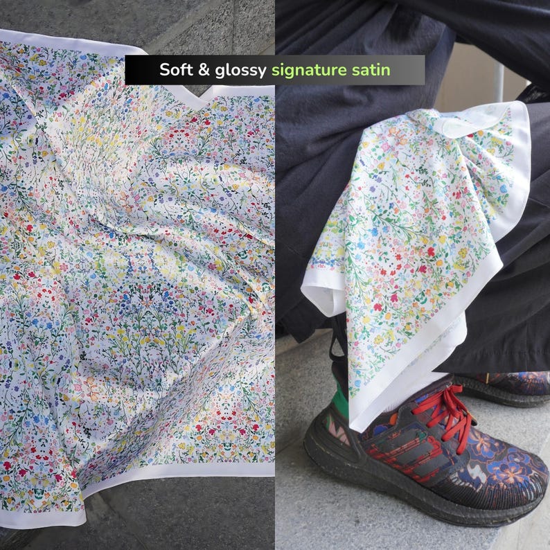 Op de afbeelding: Witte satijnen sjaal met een bloemenpatroon in blauw, geel, roze en groen. De sjaal heeft een witte rand en de tekst "Soft & glossy signature satin". De sjaal is over een been gedrapeerd.