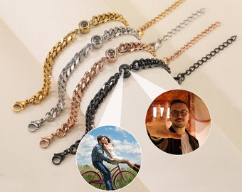 May include: Five curb chain bracelets in gold, silver, rose gold, and black. Each bracelet has a small, round charm. The image also shows a man making a frame with his hand and a couple on a bicycle.