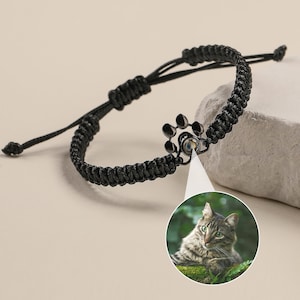 May include: Black braided bracelet with a paw print charm that reveals a photo of a tabby cat when viewed through the charm.