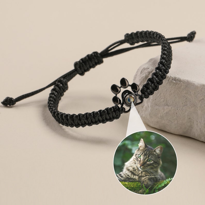 May include: Black braided bracelet with a paw print charm that reveals a photo of a tabby cat when viewed through the charm.