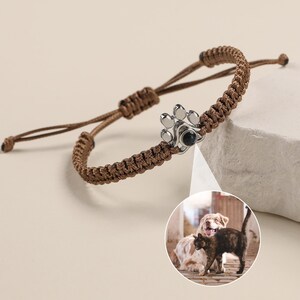 May include: A brown braided cord bracelet with a silver paw print charm. The charm has a small black photo insert, showing a dog and cat.