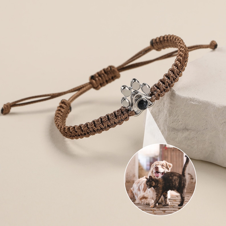 May include: A brown braided cord bracelet with a silver paw print charm. The charm has a small black photo insert, showing a dog and cat.