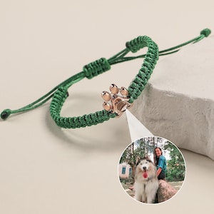 May include: A green braided bracelet with a rose gold paw print charm. The bracelet is adjustable and has a sliding knot closure.