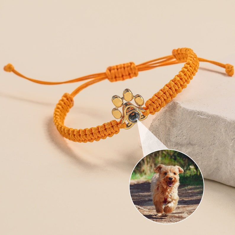 May include: Orange braided bracelet with a gold paw print charm. The charm has a small clear crystal in the center.
