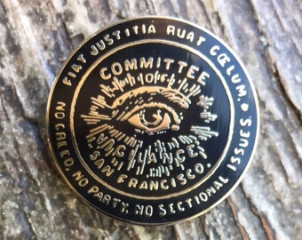 May include: A circular black and gold enamel pin featuring an eye graphic at the center. The text reads: FIAT JUSTITIA RUAT CŒLUM, COMMITTEE, VIGILANCE, SAN FRANCISCO, NO CREED, NO PARTY, NO SECTIONAL ISSUES. The pin has a metallic gold border.
