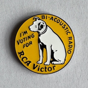 Op de afbeelding: Een ronde gele emaille speld met een witte hond met zwarte vlekken die in het midden zit. De tekst I'M VOTING FOR RCA VICTOR en BI-ACOUSTIC RADIO staat in zwarte letters rond de rand van het cirkelvormige ontwerp.
