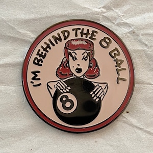 Puede incluir: Un pin de esmalte rosa y rojo con una mujer de dibujos animados sosteniendo una bola de billar negra y blanca. El texto alrededor de la mujer dice "I'm Behind The 8 Ball".
