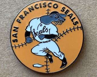 May include: A circular enamel pin featuring a tan background with black text reading SAN FRANCISCO SEALS. A seal wearing a white baseball uniform with the word seals in script on the chest is depicted in a running pose over a baseball stitch pattern.