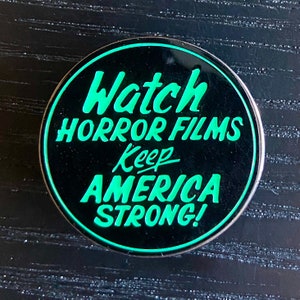 May include: A round, black pin with a silver border. The pin features the text "Watch HORROR FILMS Keep AMERICA STRONG!" in bright green, stylized lettering. The pin is set against a dark background.
