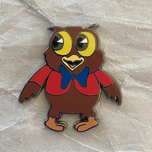 May include: A metal enamel pin featuring a brown owl character with large yellow eyes, a red jacket, and a blue bowtie. The owl has a light-colored beak and tan feet, with black outlines defining the shape and features of the cartoon-style bird.
