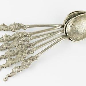 Könnte beinhalten: Ein Set aus fünf silberfarbenen Metalllöffeln mit kunstvoll gestalteten drachenförmigen Griffen und Laffen, die mit detaillierten Blumenmustern geprägt sind. Die Löffel sind fächerförmig angeordnet und zeigen die strukturierte Metallverarbeitung.