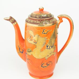 Könnte beinhalten: Orangefarbene Keramik-Teekanne mit goldenem Drachendesign. Die Teekanne hat einen Deckel mit goldenem Knauf und einen Griff mit goldenem Besatz. Der Drache ist goldfarben mit blauen Akzenten. Ausguss und Deckel haben Zierleisten.