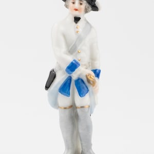 Puede incluir: Figura de porcelana de un hombre con atuendo del siglo XVIII. Viste abrigo blanco, medias grises y sombrero negro con ala blanca. Detalles azules adornan el abrigo y la faja. La figura se alza sobre una base rectangular con ribete dorado.