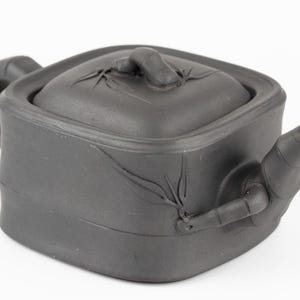 May include: A dark grey ceramic teapot with a square body and a lid. The teapot has a curved handle and a spout that resembles bamboo. The lid features a small handle and decorative bamboo leaf details. The teapot is set against a white background.