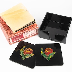 Puede incluir: Un juego de posavasos lacados en negro con un diseño floral, junto con un soporte negro a juego y una caja decorativa. Los posavasos presentan una flor roja y amarilla con hojas verdes. La caja es roja con un diseño.